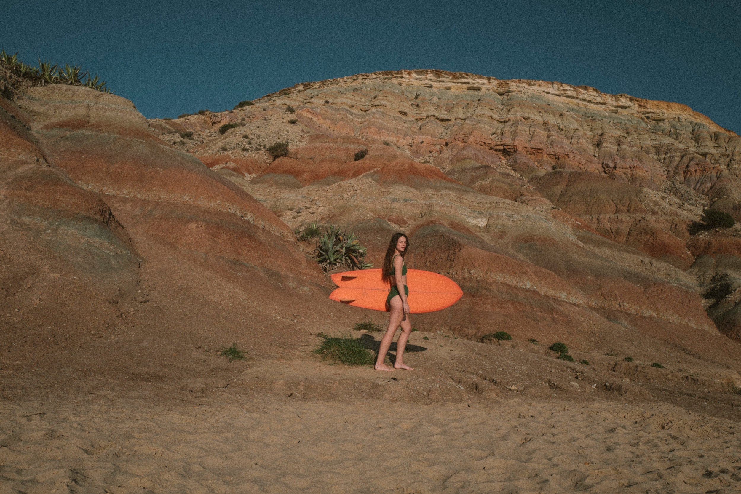 Person holding an orange surfboard in a desert landscape with red rock formations.